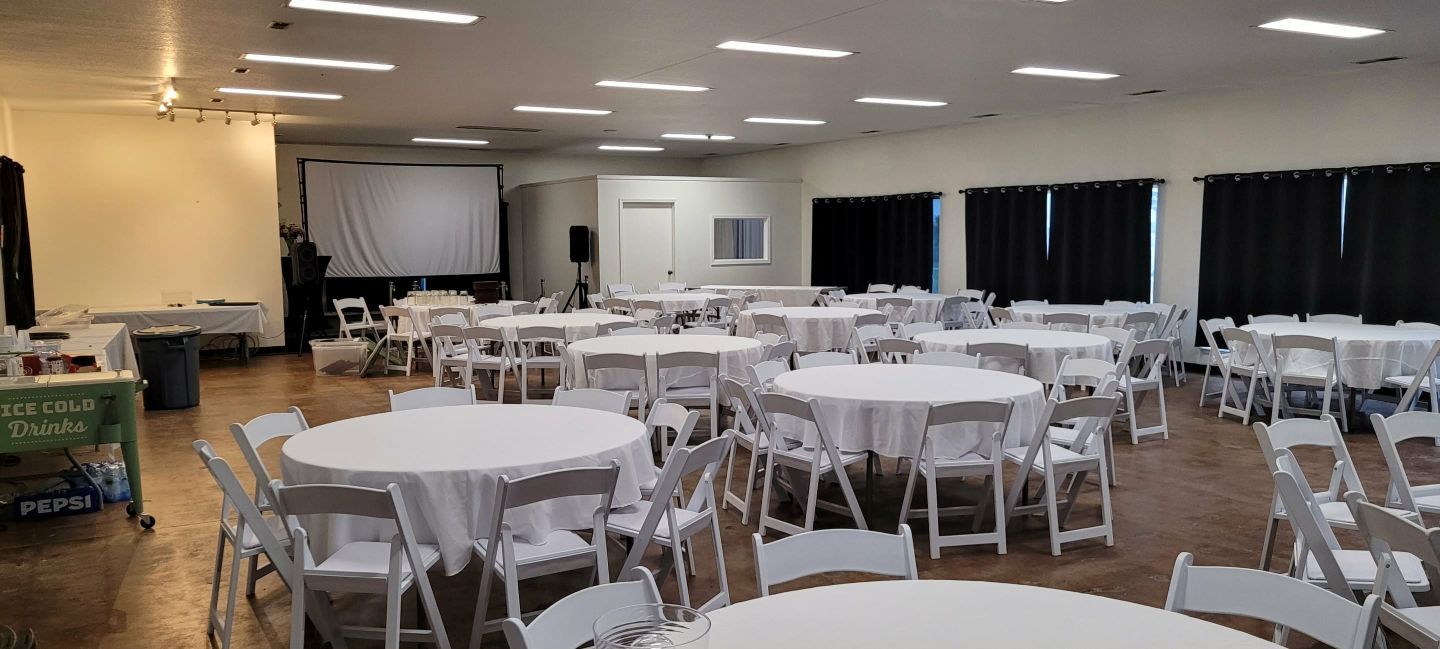 A large room set up for an event with round tables covered in white tablecloths and white chairs.