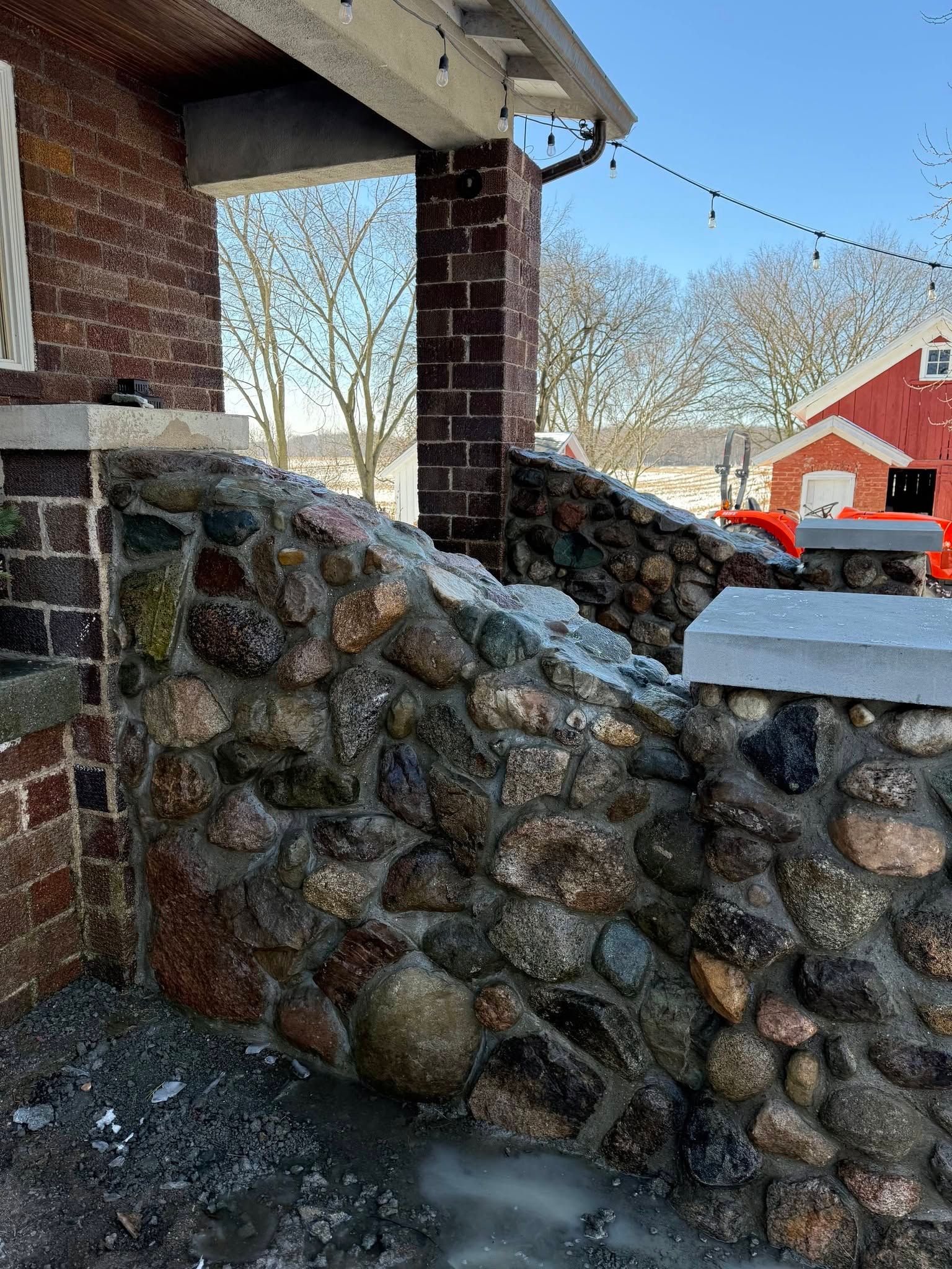 Snow-covered stone wall on a porch with a brick pillar and a red house in the background.