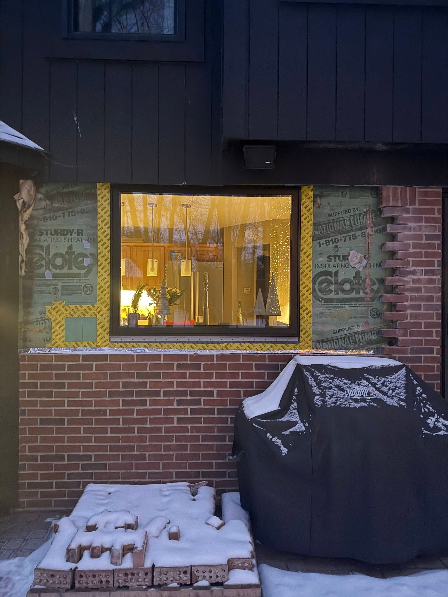 Brick patio with snow, a lit window, and a covered grill against a dark house exterior.