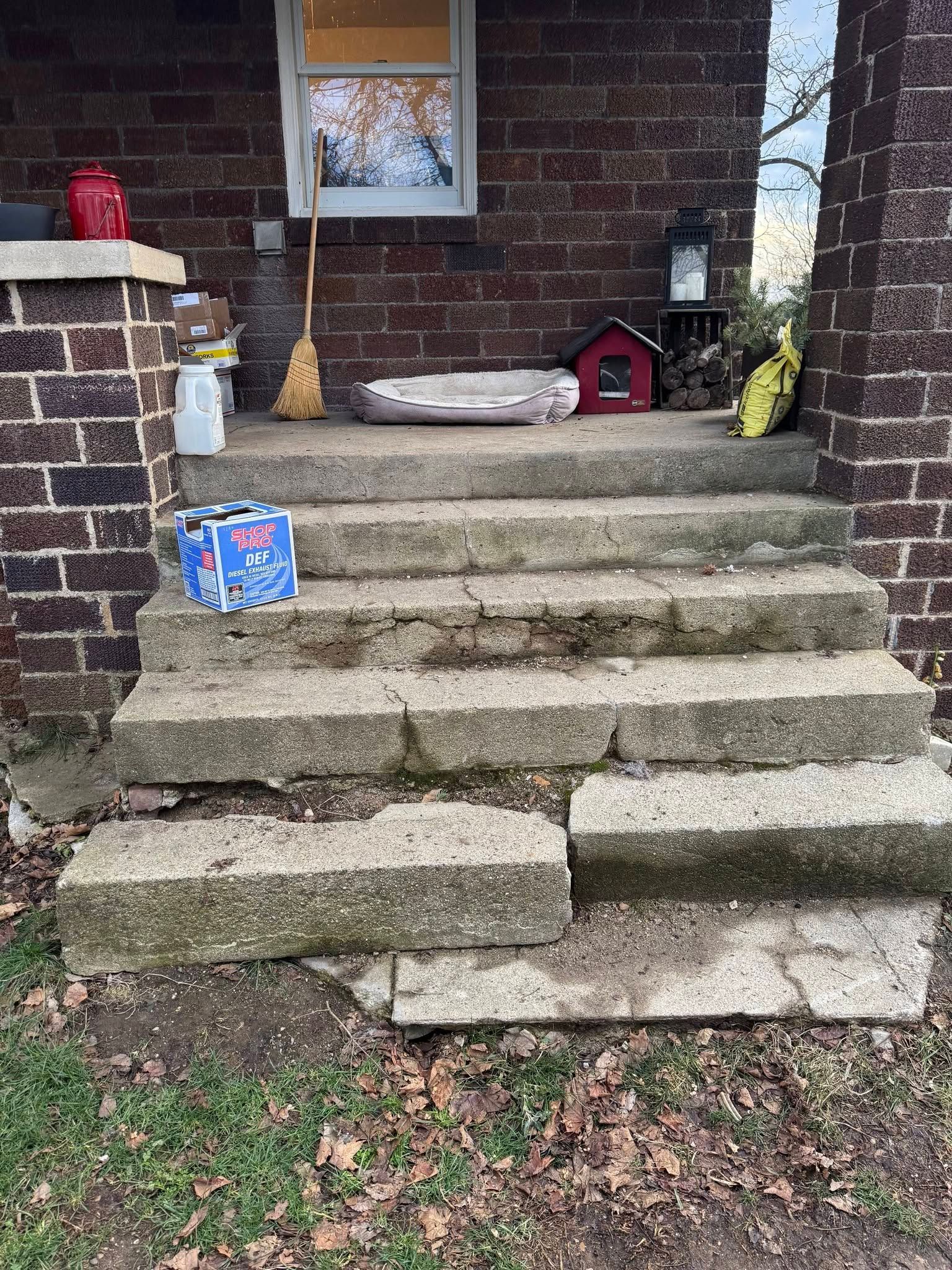Broken concrete steps leading to a brick porch with a red birdhouse and yard tools nearby