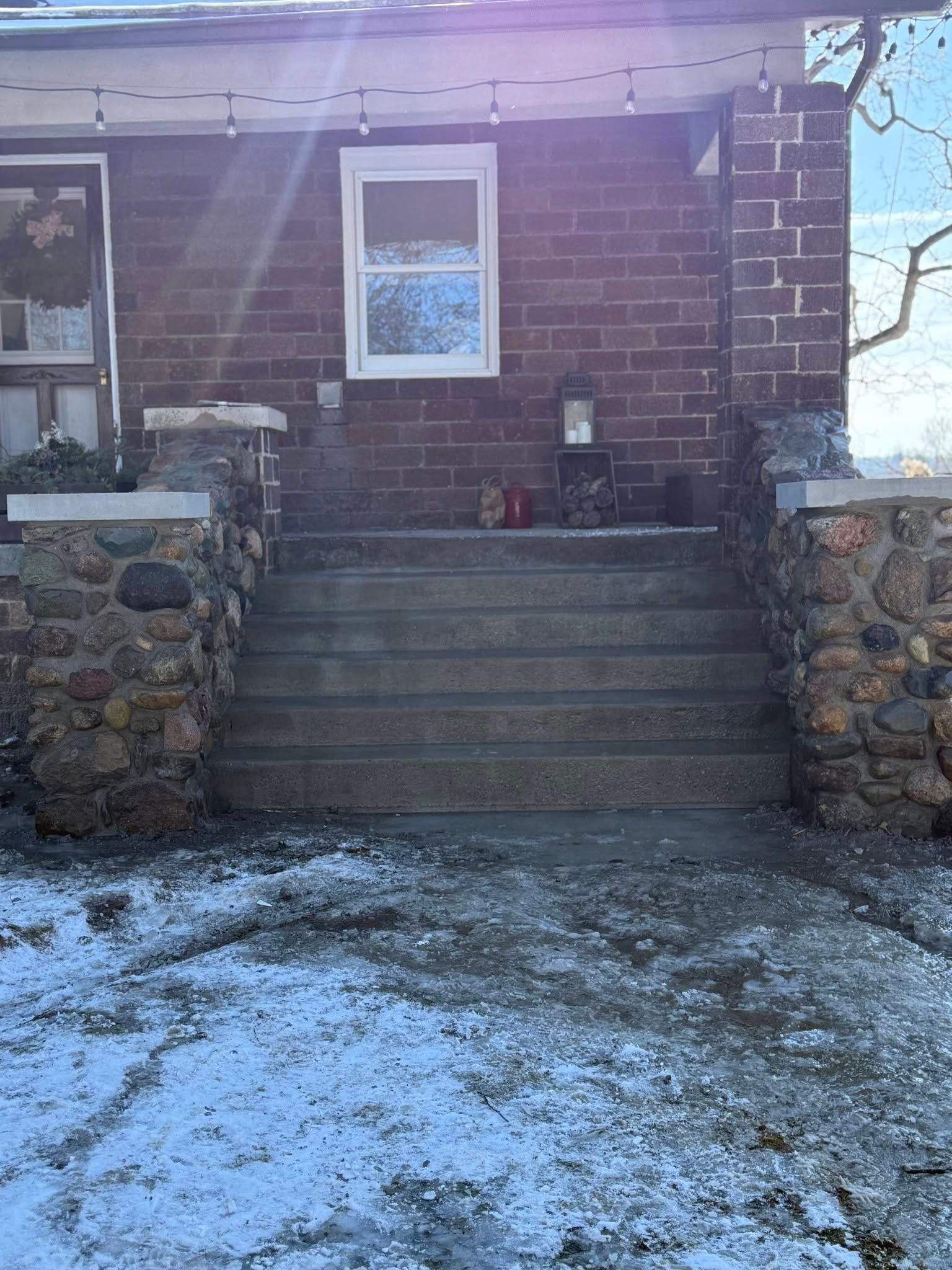 Snowy front steps leading to a brick house entrance with a white-framed window and stone side walls