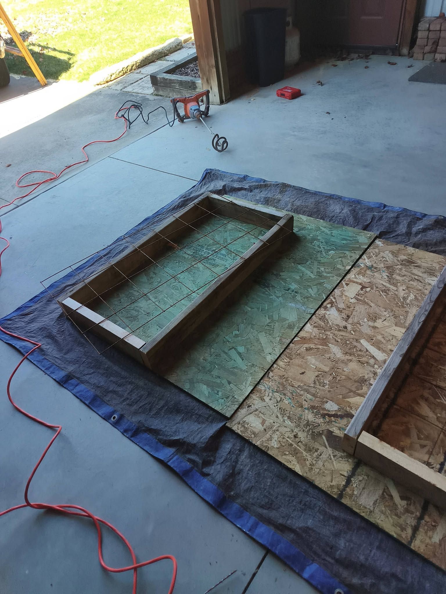 Construction area with a rectangular floor opening framed by boards and tarp on a concrete slab.