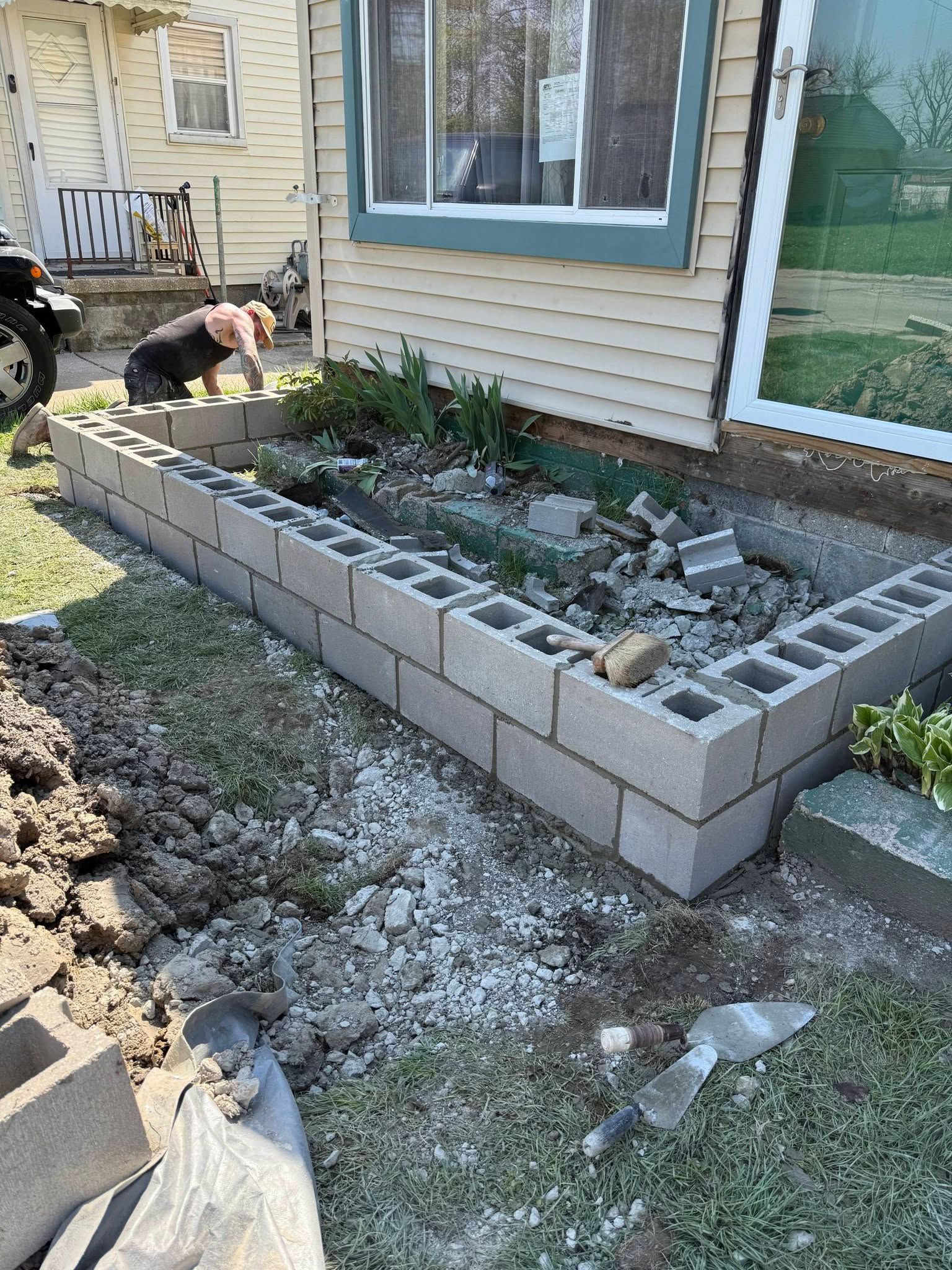Outdoor yard project with cinder block raised garden bed beside a house, partially filled with soil and plants