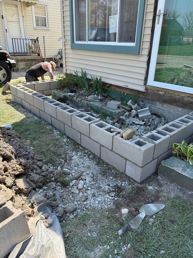 Person building a raised garden bed beside a house, with cinder blocks and soil in a yard
