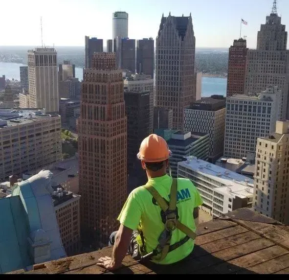 Worker in orange hard hat and green shirt sits on rooftop overlooking a city skyline.