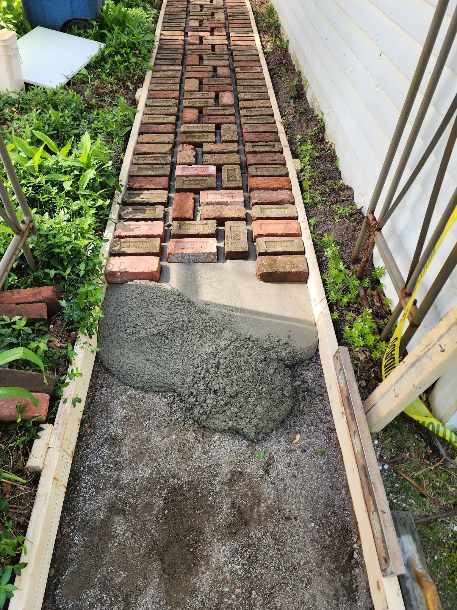 Narrow garden path with scattered bricks and a patch of wet concrete between plants and a fence