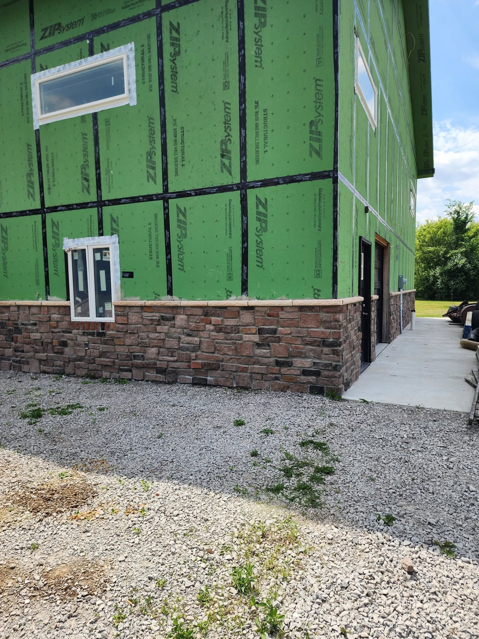 Partially built green house with exposed wrap, brick foundation, and gravel driveway beside a sidewalk