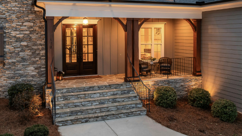Front porch at dusk with stone steps, warm porch lights, and wicker chairs beside a house entrance