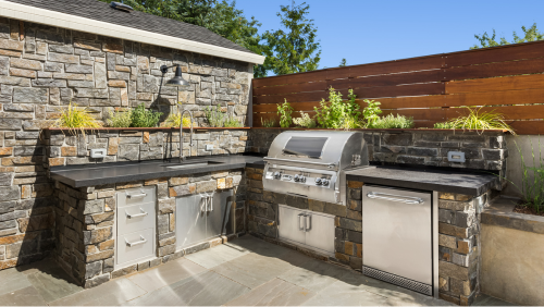 Stone outdoor kitchen with built-in grill, sink, cabinets, and countertop against a stone wall and wood fence