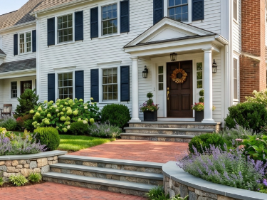 Front entrance of a white house with brick steps, black shutters, potted plants, and a landscaped garden.