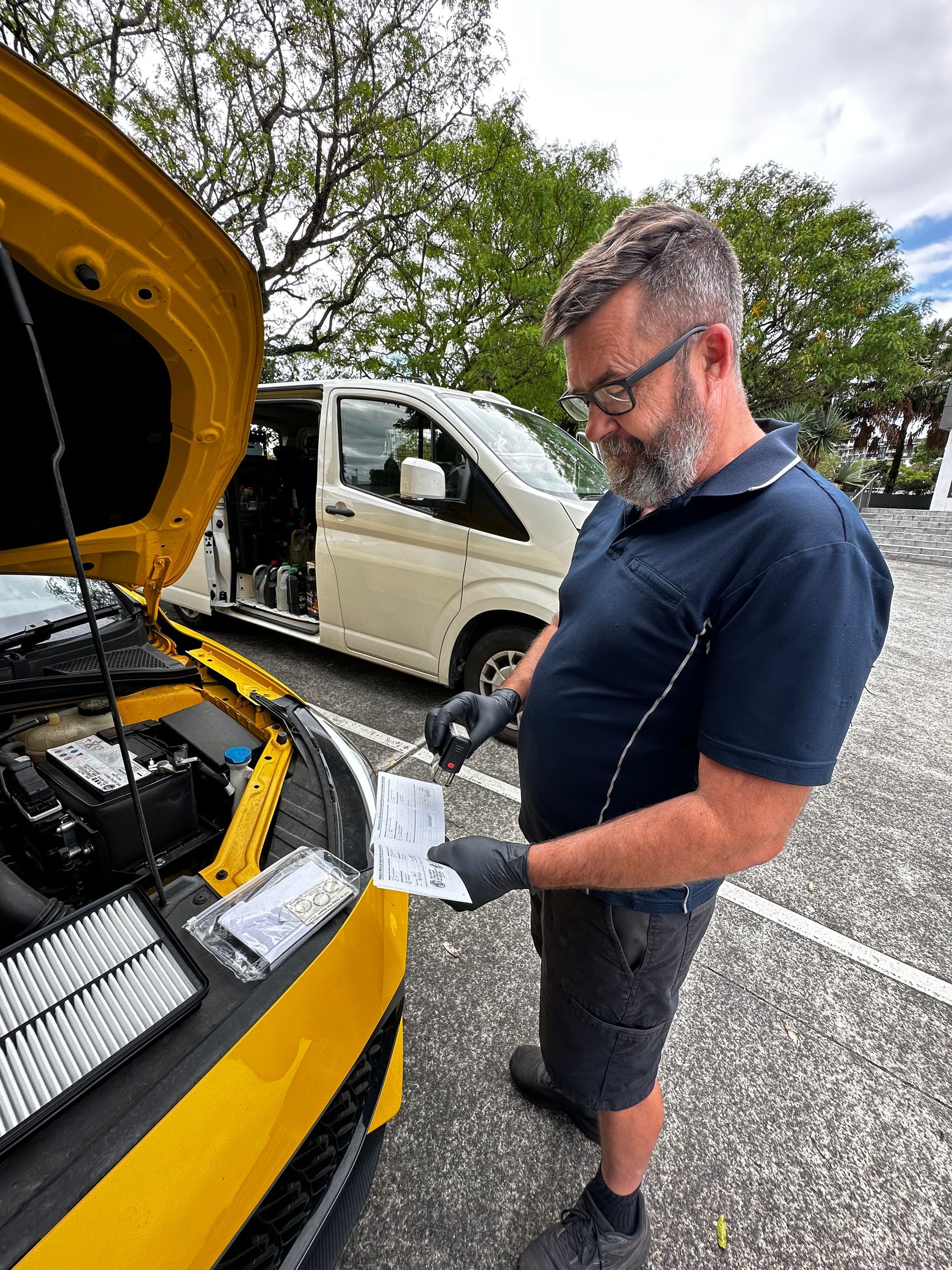 Man checking a yellow car’s engine bay while holding a part, with a white van and trees in the background — Calvin's Mobile Mechanics In Elanora, QLD
