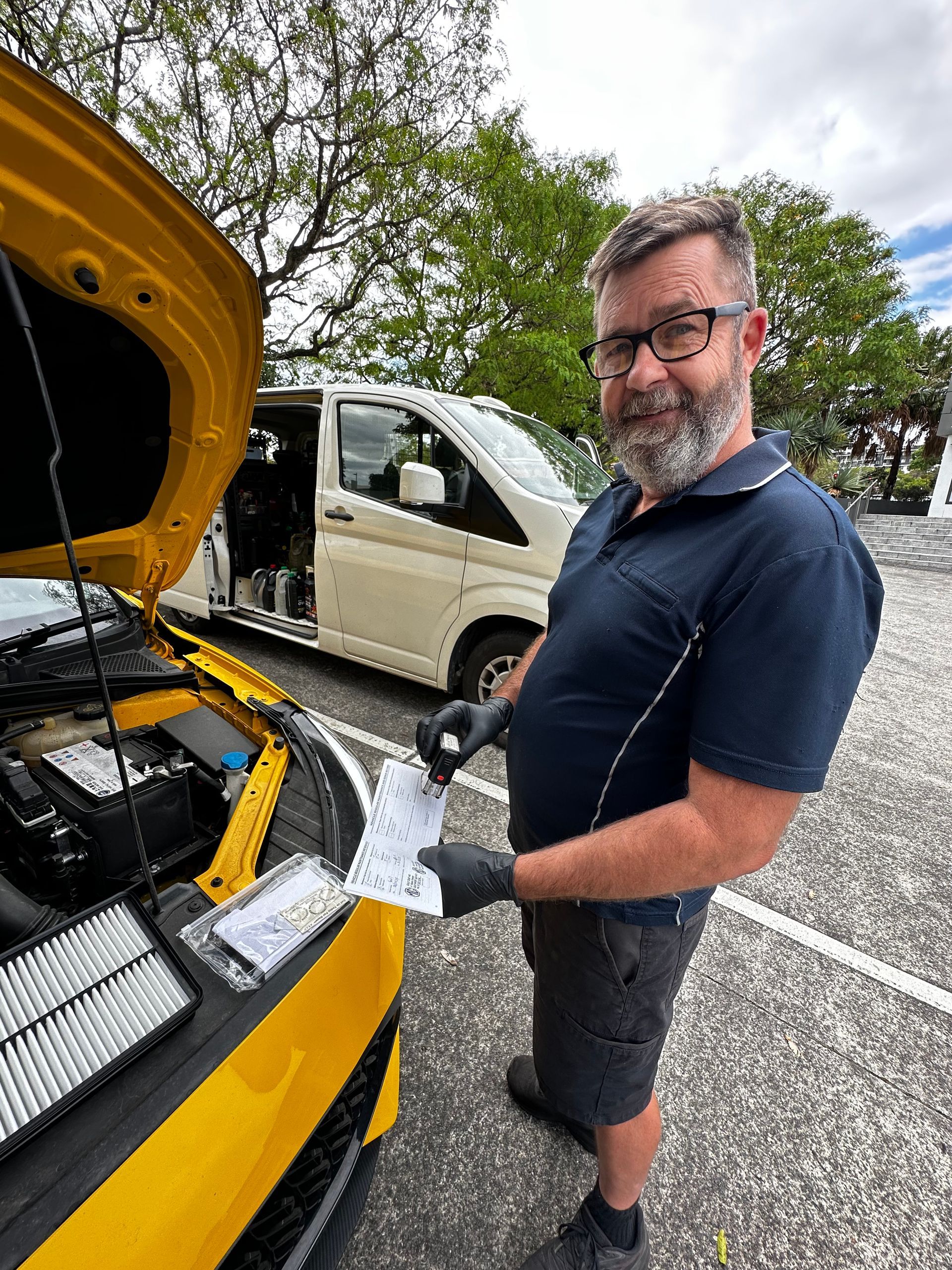 Man in a navy shirt stands beside a yellow car with the hood open, inspecting the engine in a parking lot. — Calvin's Mobile Mechanics In Elanora, QLD