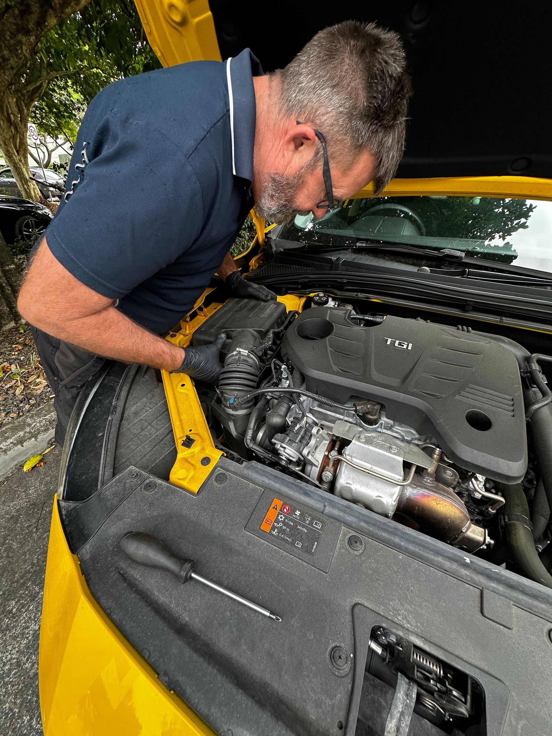 Mechanic inspecting a car engine under an open hood, wearing gloves and leaning over the engine bay. — Calvin's Mobile Mechanics In Elanora, QLD