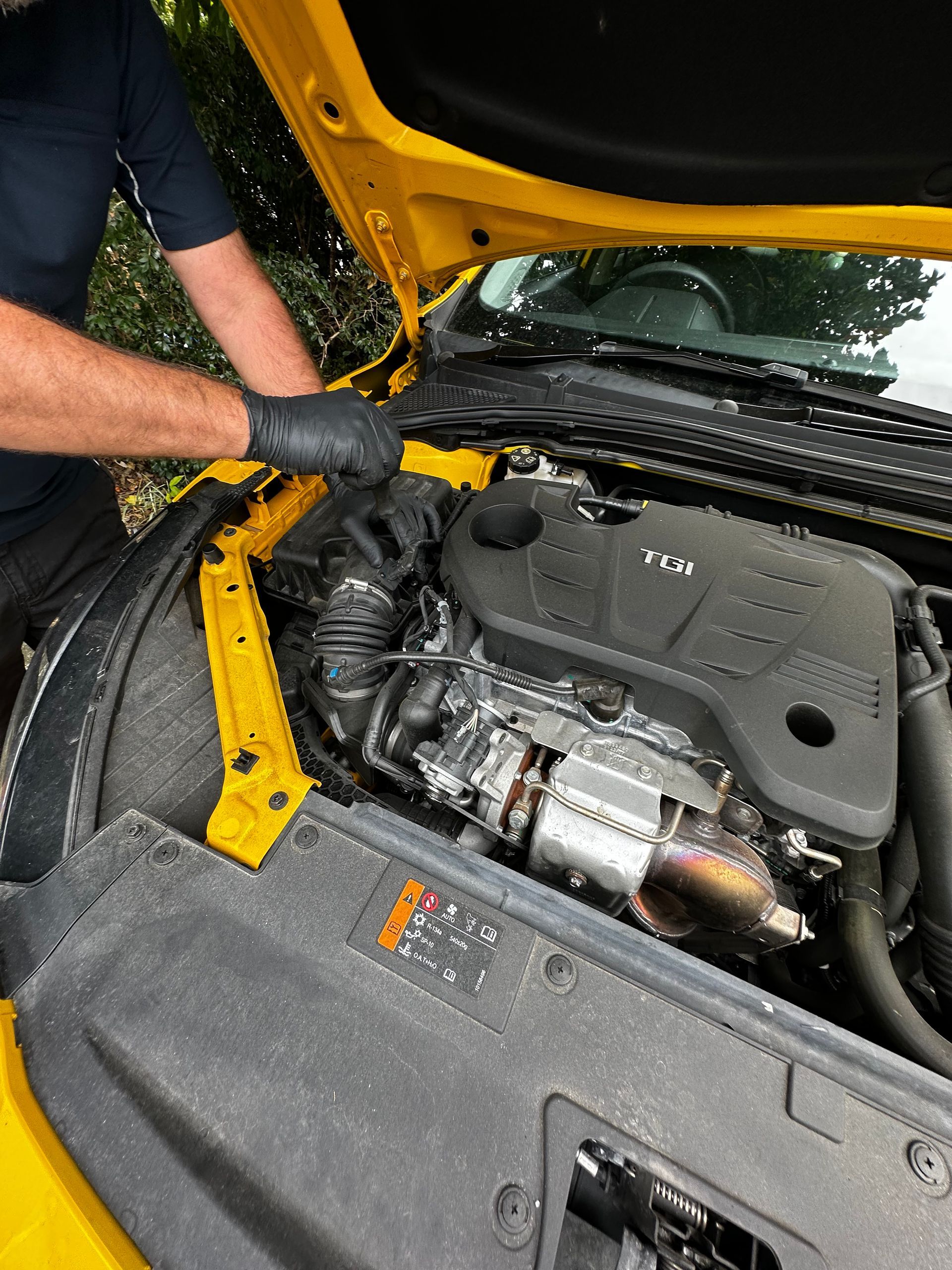 Mechanic working under a yellow car hood, inspecting the engine bay with a gloved hand — Calvin's Mobile Mechanics In Palm Beach, QLD