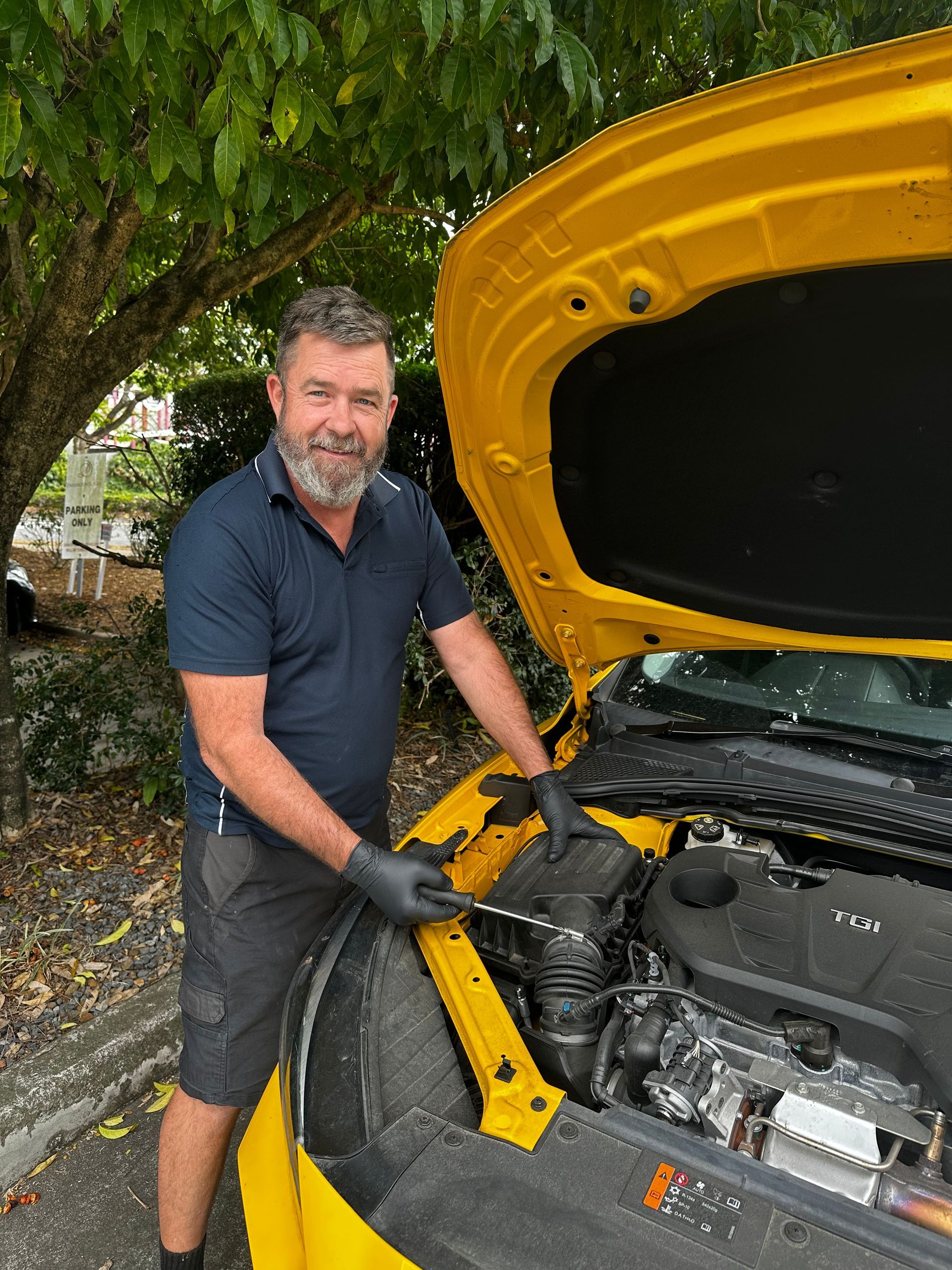 Man in a navy shirt and gloves working under the open hood of a yellow car outdoors — Calvin's Mobile Mechanics In Elanora, QLD