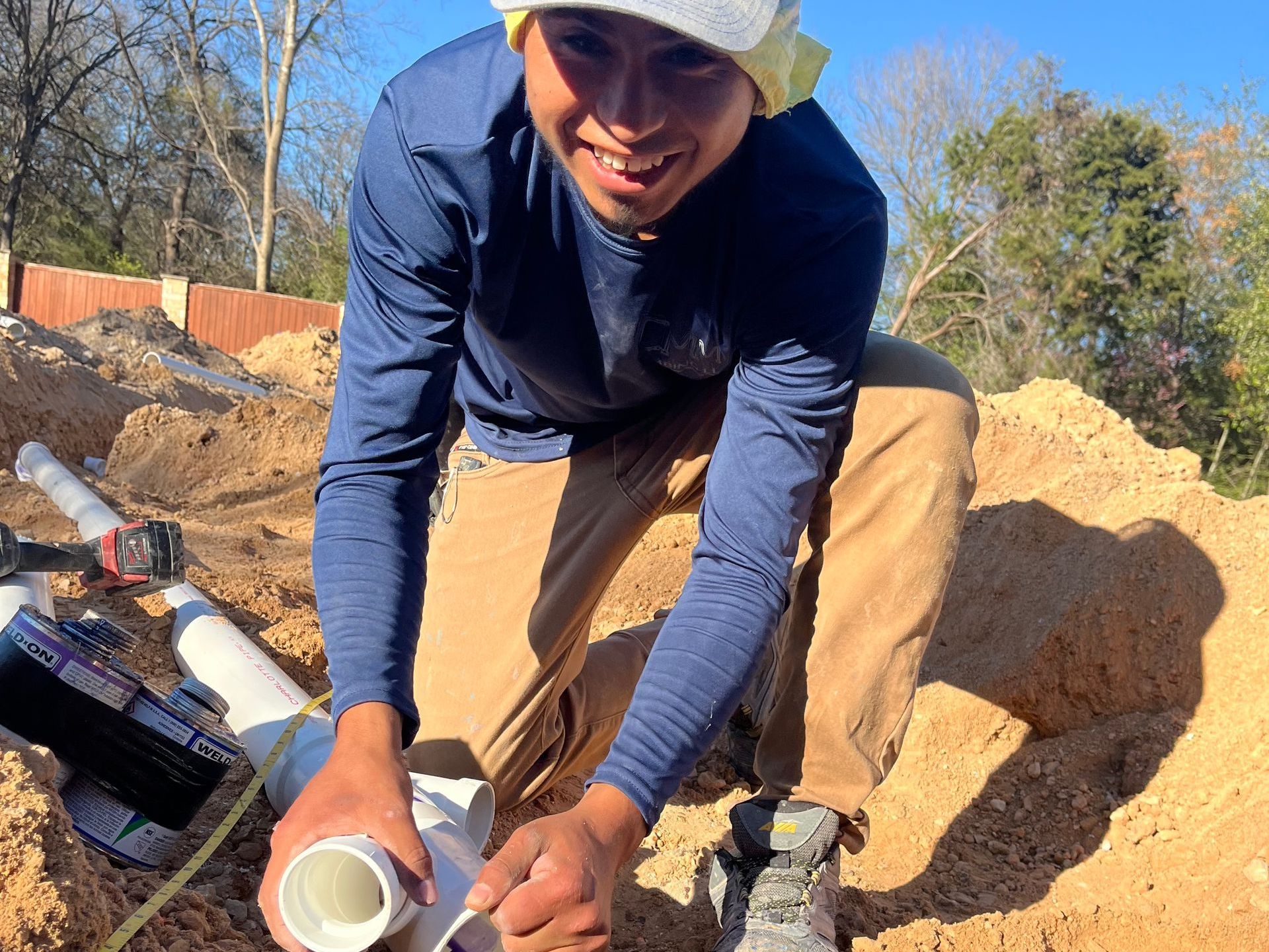 Man working on plumbing outdoors, smiling. He holds white pipes surrounded by dirt.