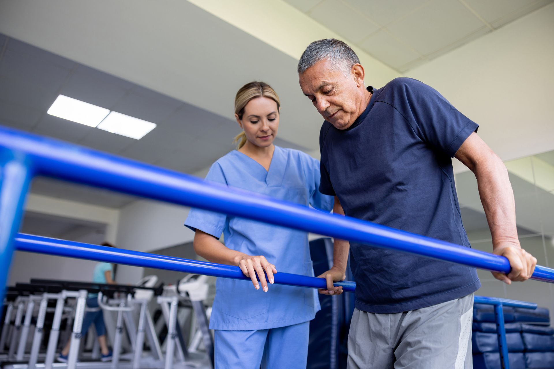 Therapist assists a person walking between blue parallel bars in a rehabilitation gym.