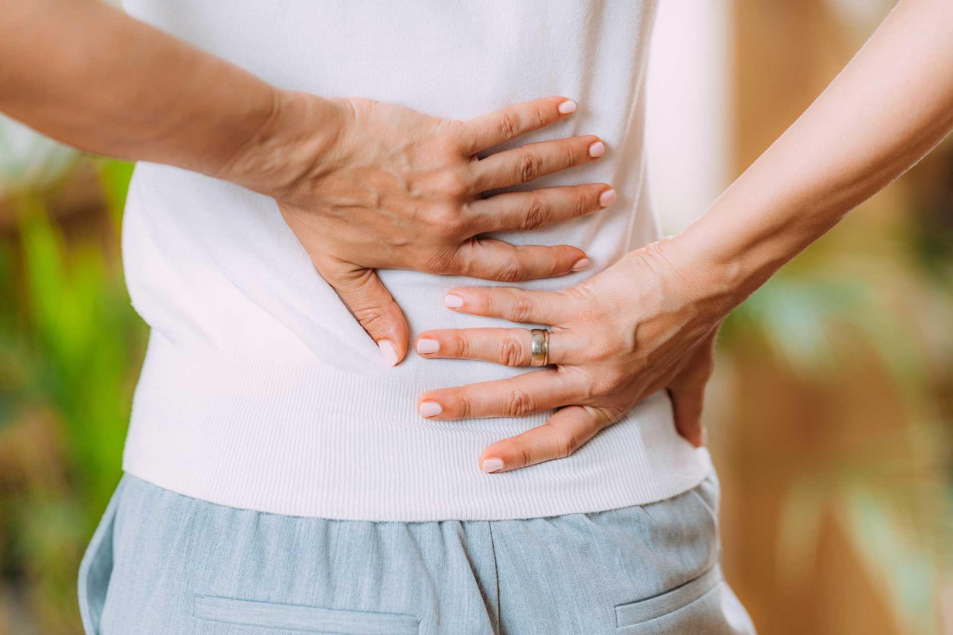 Woman with sciatic nerve pain in her lower back. Woman holding her lower back in pain; white shirt, light blue pants, hands visible.