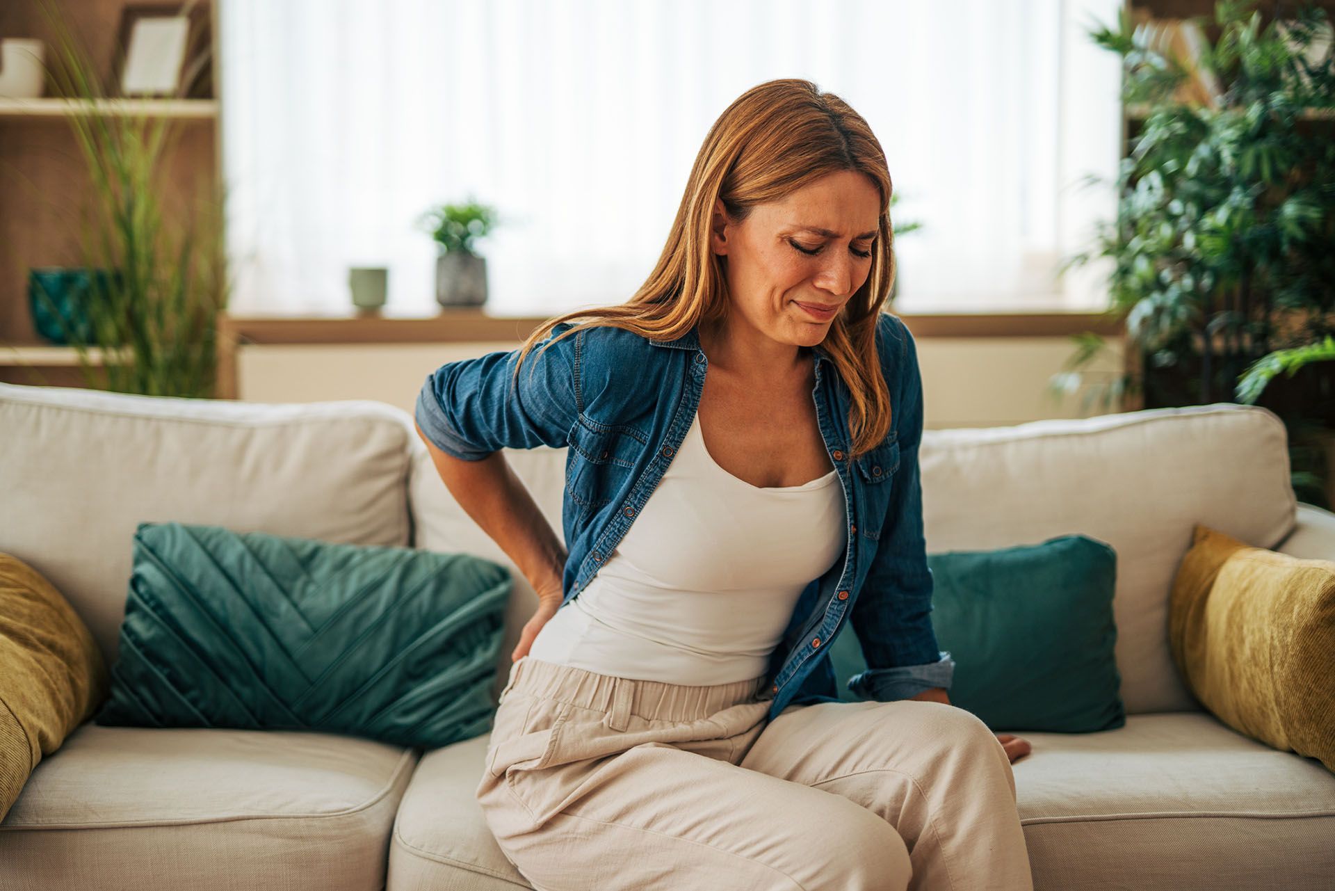 A woman is suffering from back pain while sitting on a sofa. Woman sitting on a couch, clutching her lower back in pain.
