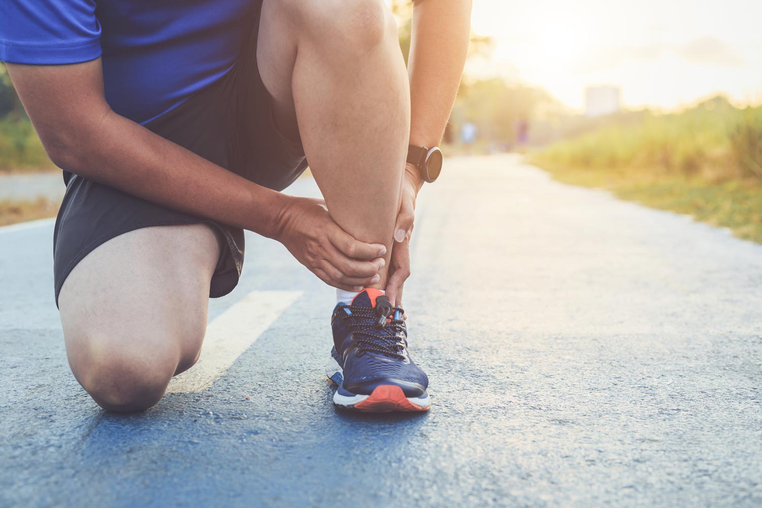 Athlete Suffering From Joint Injury — McAllen, TX — Texas Rehab Center Person holding their ankle, kneeling on a blue running track, presumably injured, sunlight.