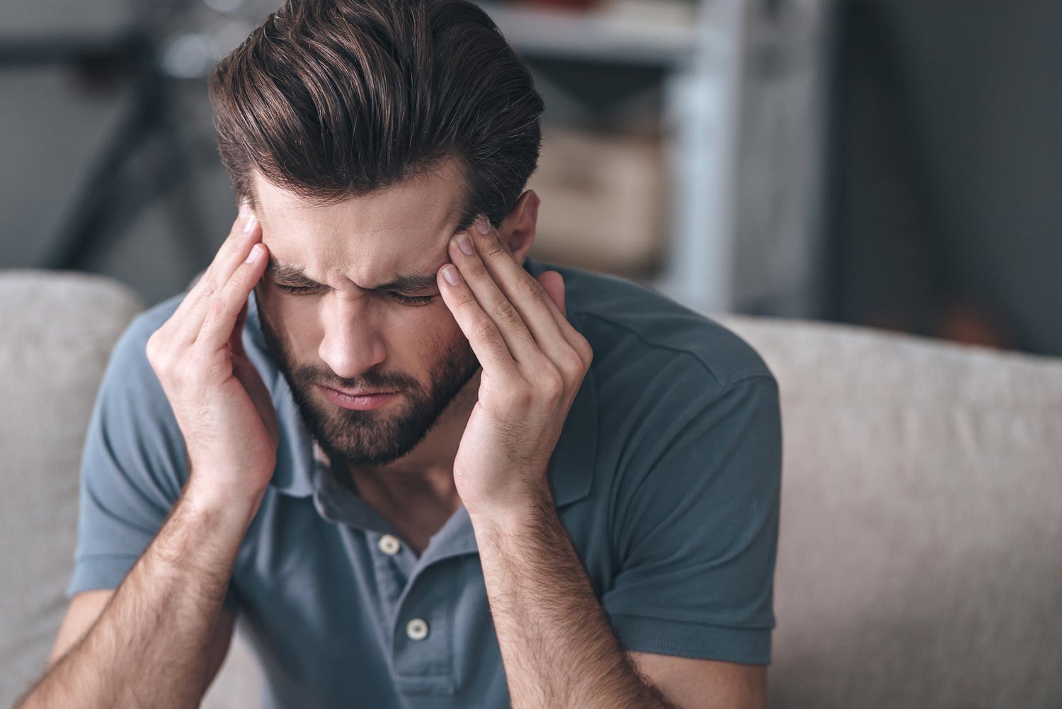 Man with hands on temples, seated indoors, appearing to have a headache.