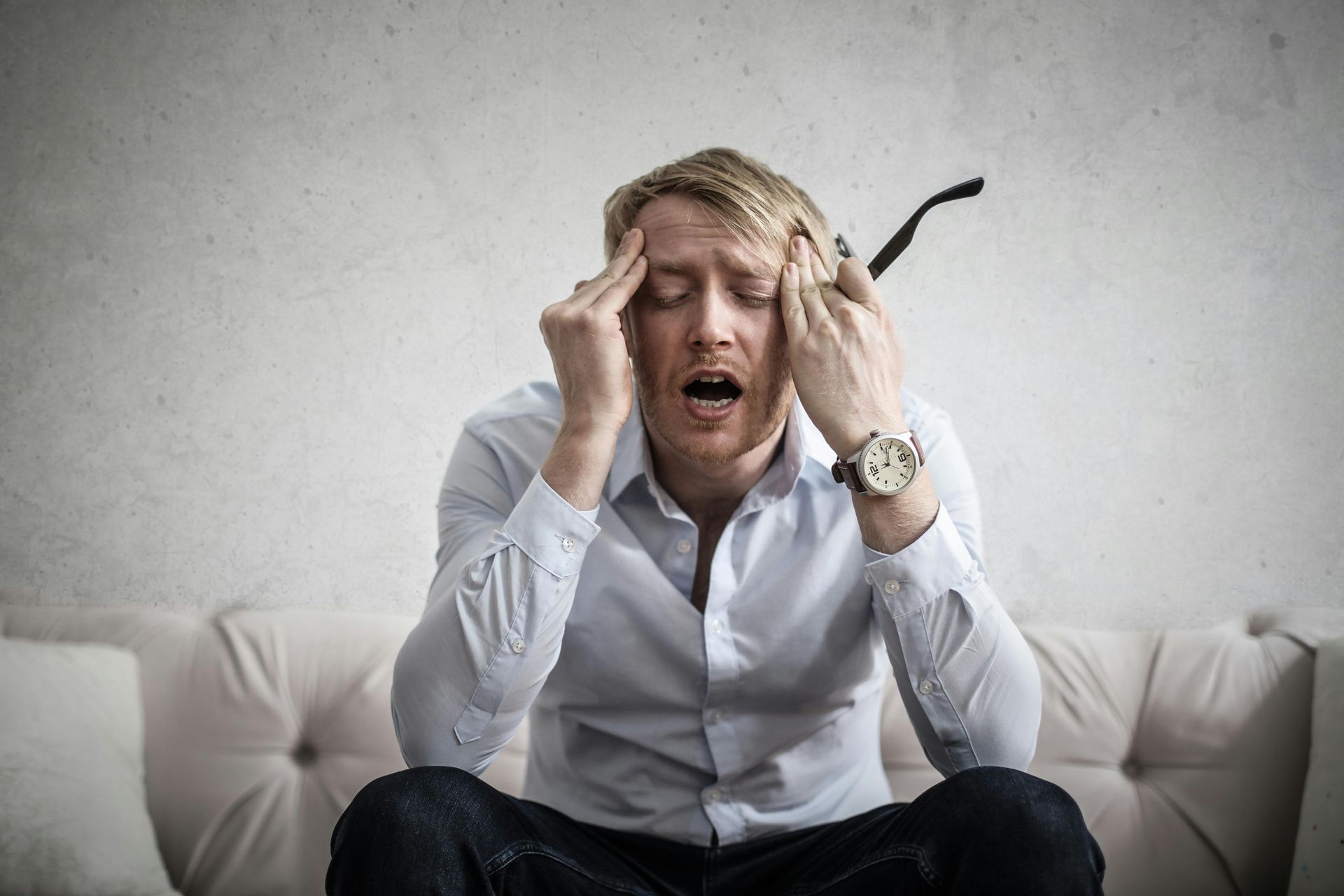 Man with hands on head, eyes closed, mouth open in distress; sitting on a couch.