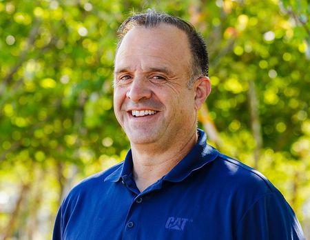 Smiling man in a blue CAT shirt, outdoors with green foliage in the background.
