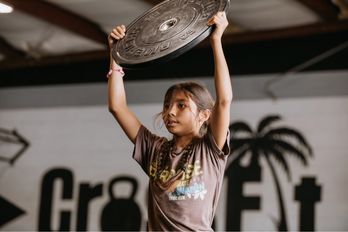 Child lifting a weight plate overhead in a gym with a blurred CrossFit sign in the background