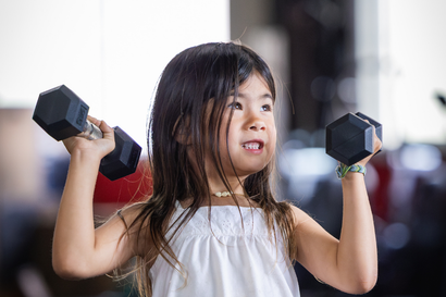 Child lifting dumbbells in a gym, wearing a white top and looking to the side.