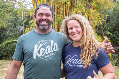 Smiling couple in matching Keala shirts standing outdoors in front of greenery