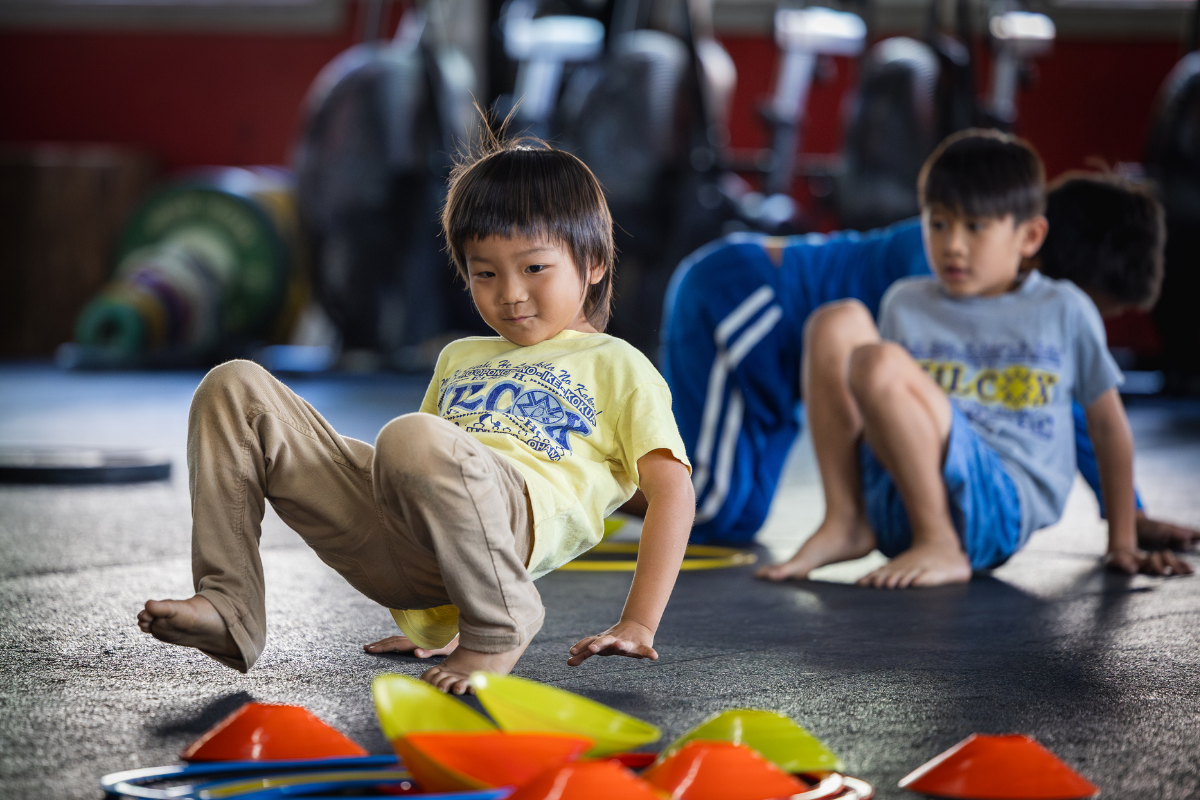 Two children exercising on mats in a gym, with colorful cones in the foreground.