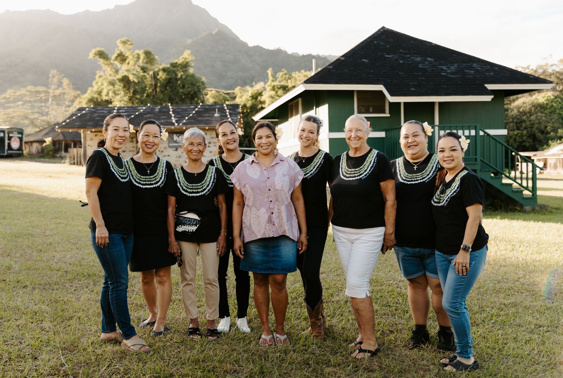 Eight women stand together outdoors in front of a green building and mountain backdrop.