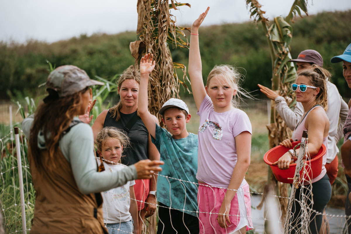 Children and adults in a field holding corn stalks, smiling and raising their hands