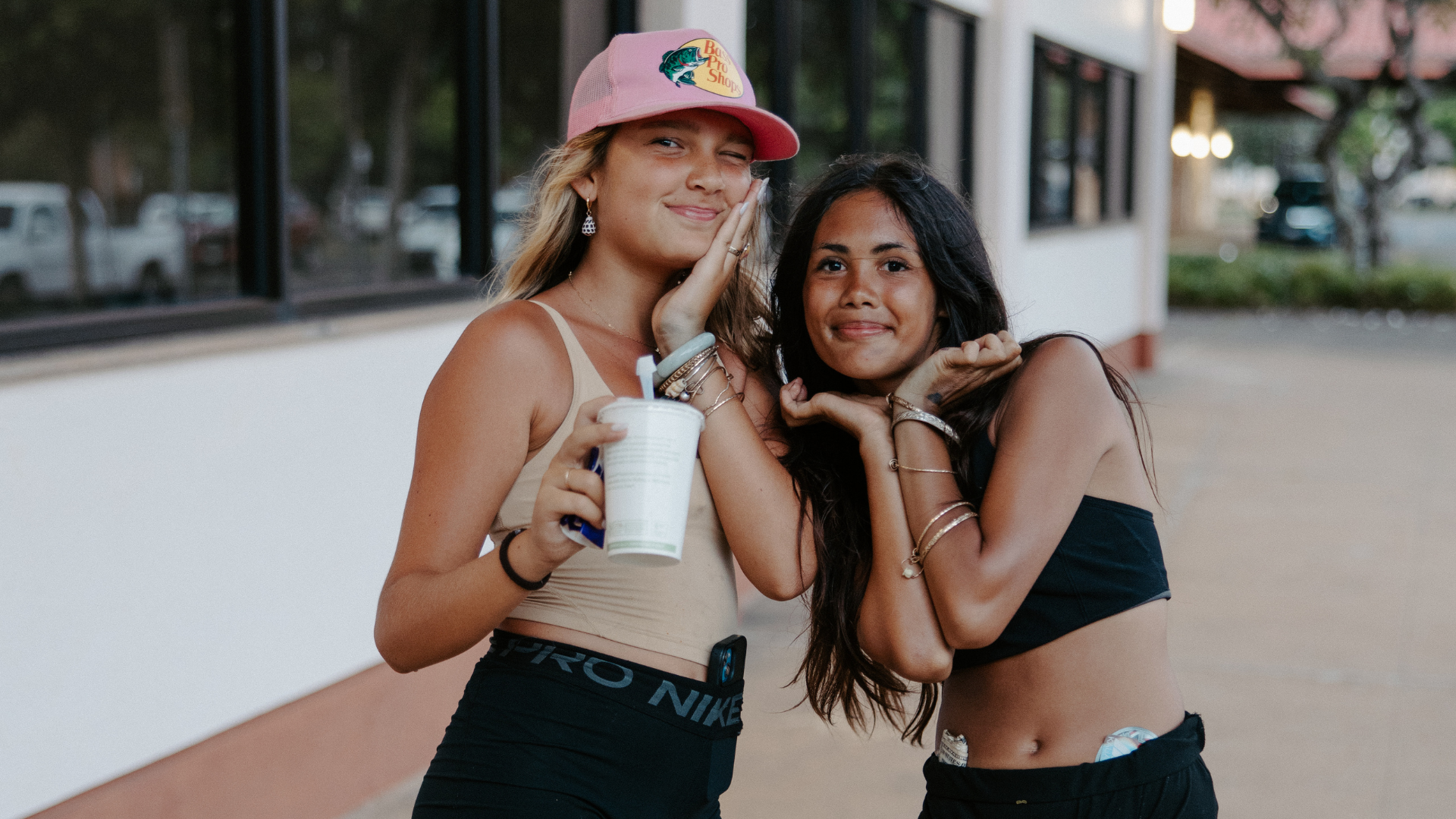 Two friends posing outdoors, one holding a drink, both smiling and leaning together in casual outfits.