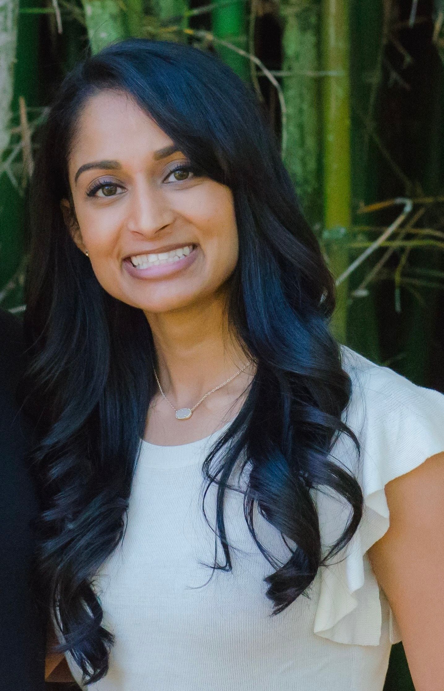 A woman with long black hair is smiling in front of a bamboo forest.