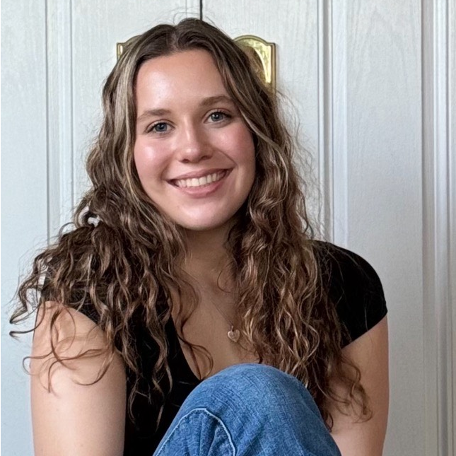 A woman with curly hair is sitting on the floor in front of a door and smiling.