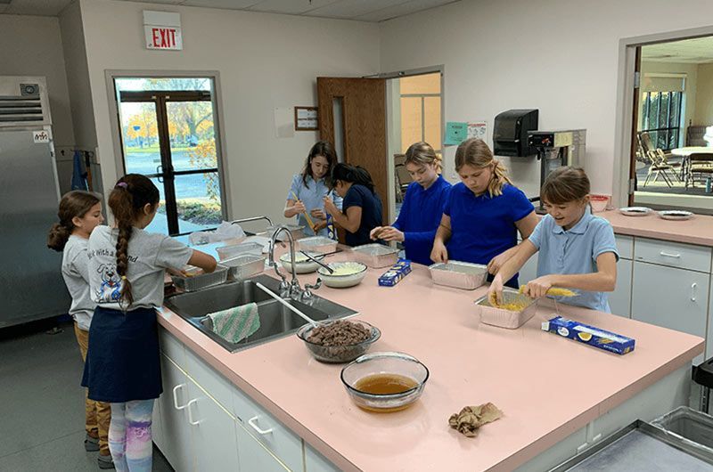 Children in a kitchen, preparing food at a pink countertop, near sinks and bowls. Tuition & Registration | Immanuel Lutheran School | Belvidere, IL