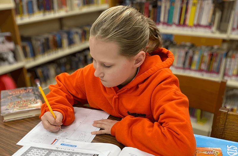 Girl in an orange hoodie, writing with a pencil at a table in a library. Discipleship | Immanuel Lutheran School | Belvidere, IL