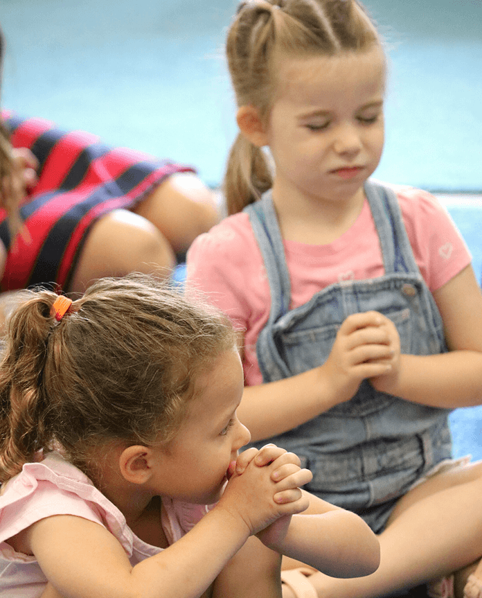 Two young girls sitting on floor, one looking at foot, other with eyes closed, hands clasped. Discipleship | Immanuel Lutheran School | Belvidere, IL