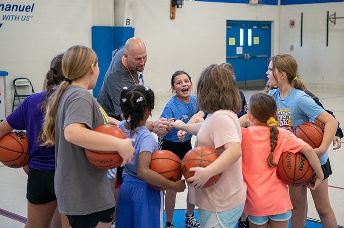 Children and a coach huddle with basketballs in a gym, smiling and laughing.