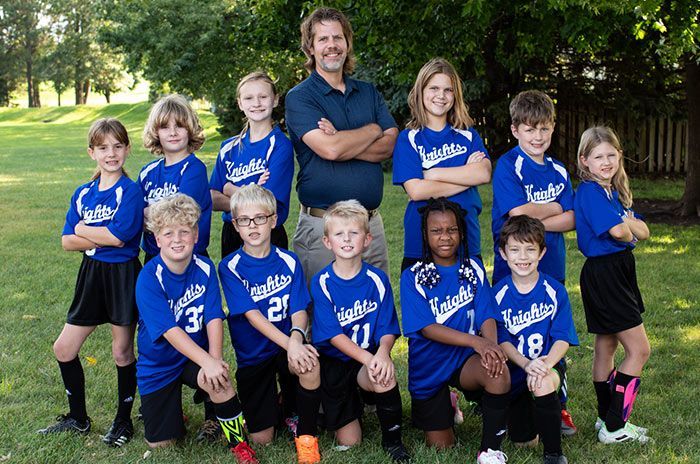 Youth soccer team in blue jerseys, posing on a green field with a coach in the background.