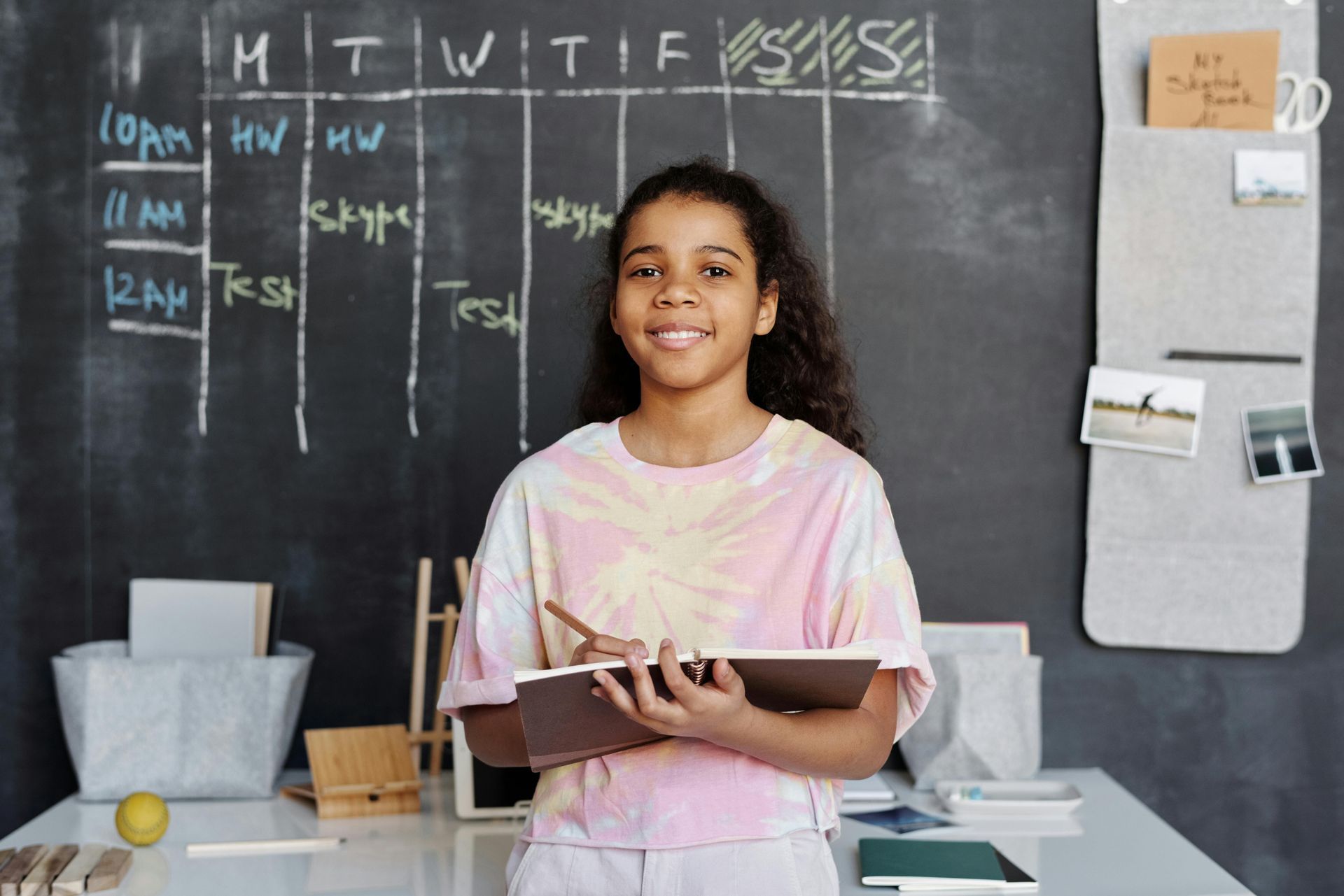Girl standing at desk with schedule chart on chalkboard behind her, holding notebook and pencil. The Immanuel Difference | Immanuel Lutheran School | Belvidere, IL