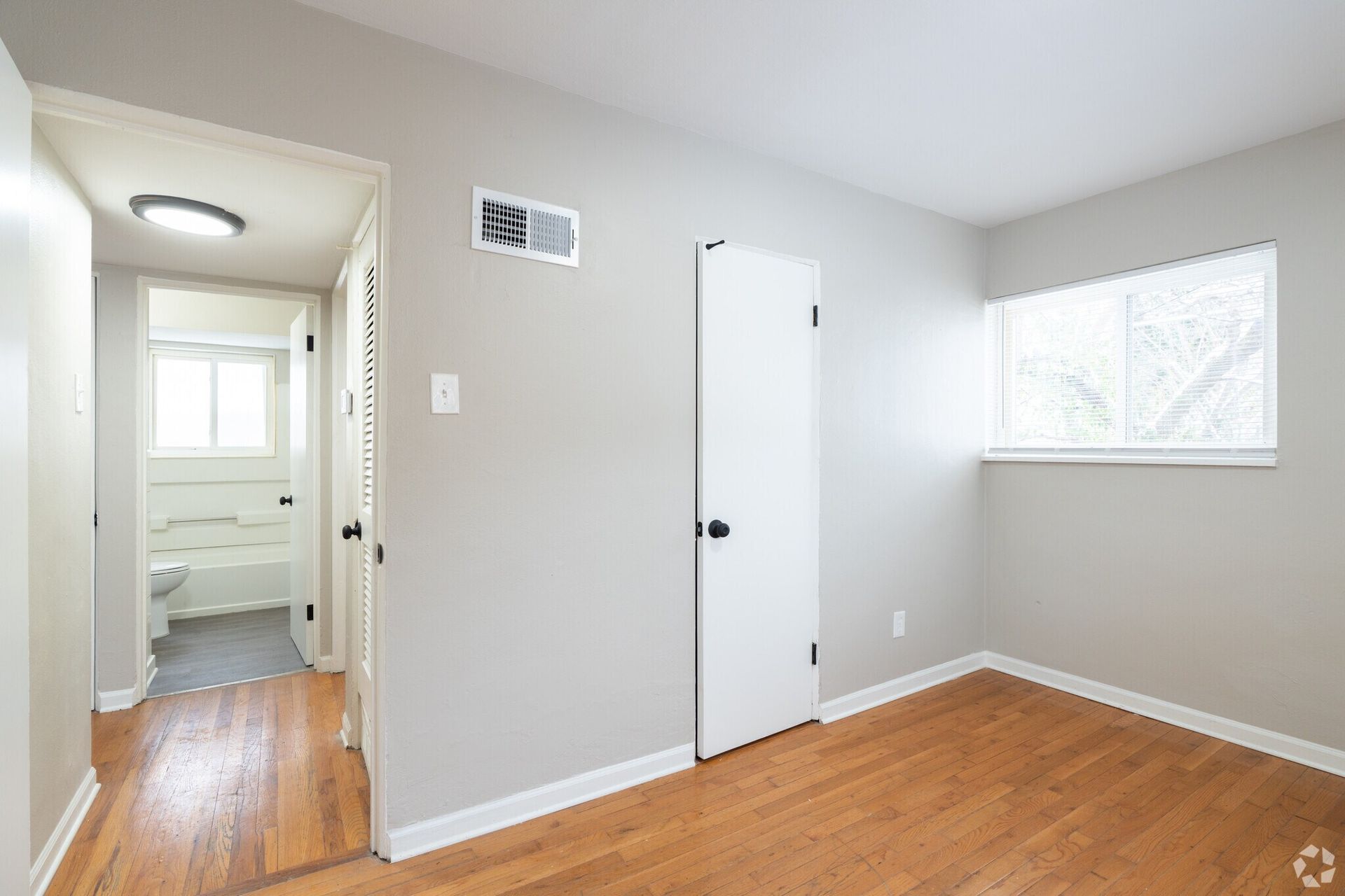 Empty beige bedroom with hardwood floors, white closet door, window, and open doorway to a bathroom.