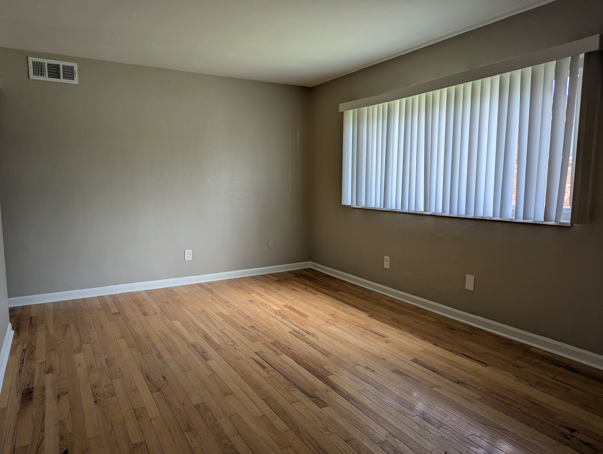 Empty beige living room with hardwood floor and a large window with vertical blinds