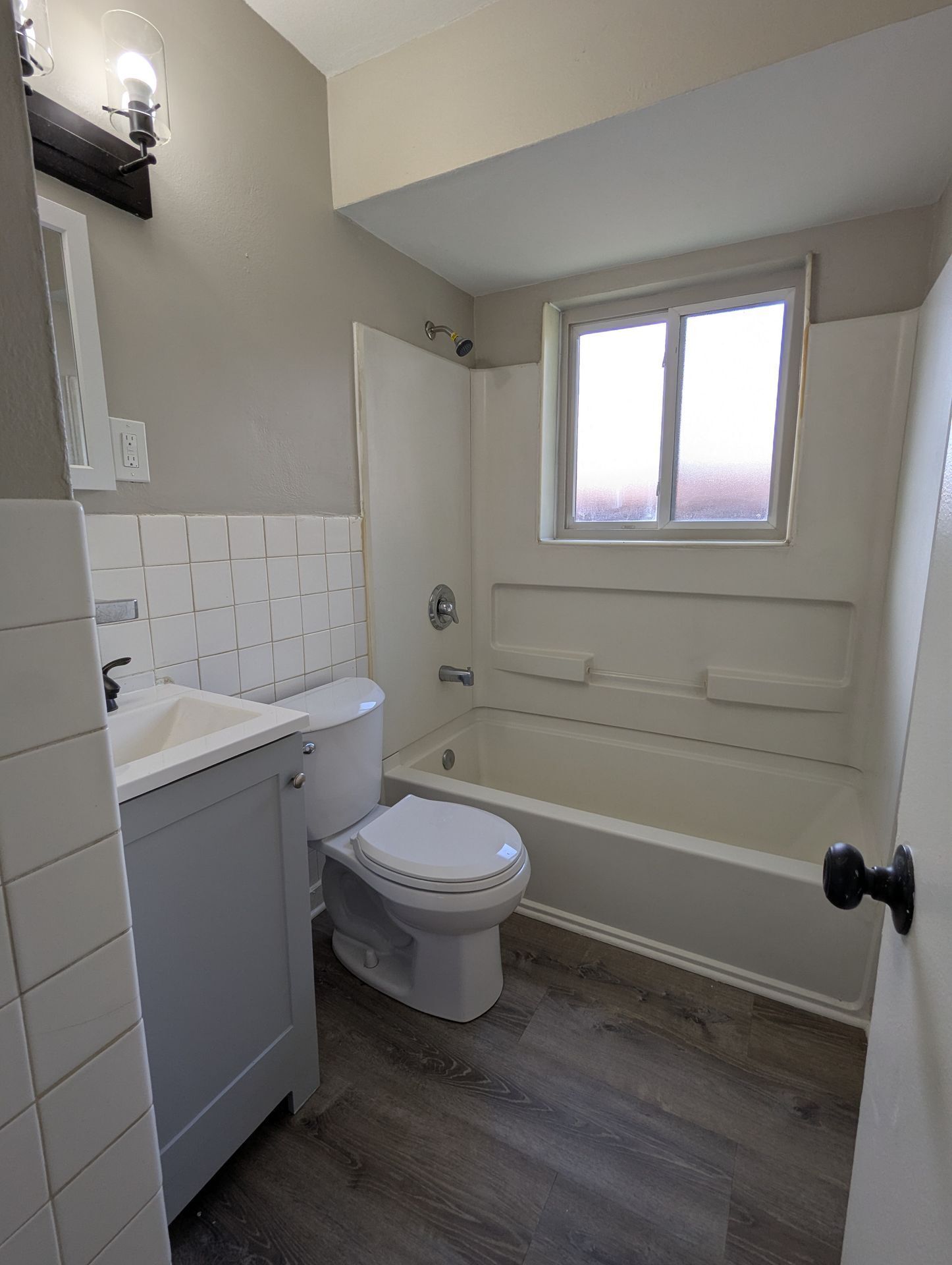 Small bathroom with toilet, vanity, bathtub, and frosted window; white tile and gray wood-look floor.
