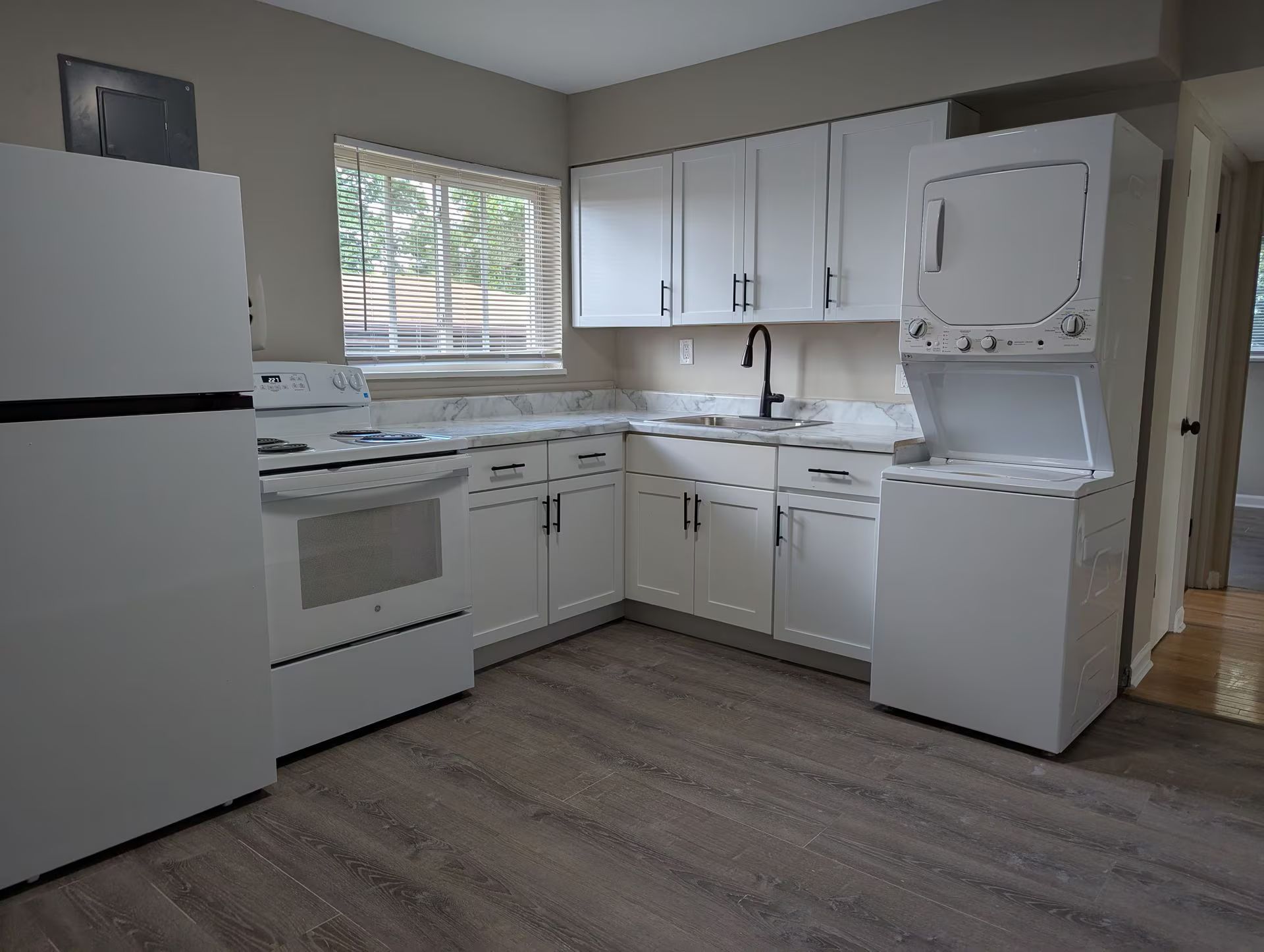 Small white kitchen with stove, fridge, cabinets, sink, window, and stacked washer and dryer.