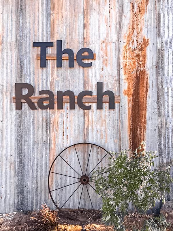 “The Ranch” sign on weathered wood wall with a wagon wheel and shrub below