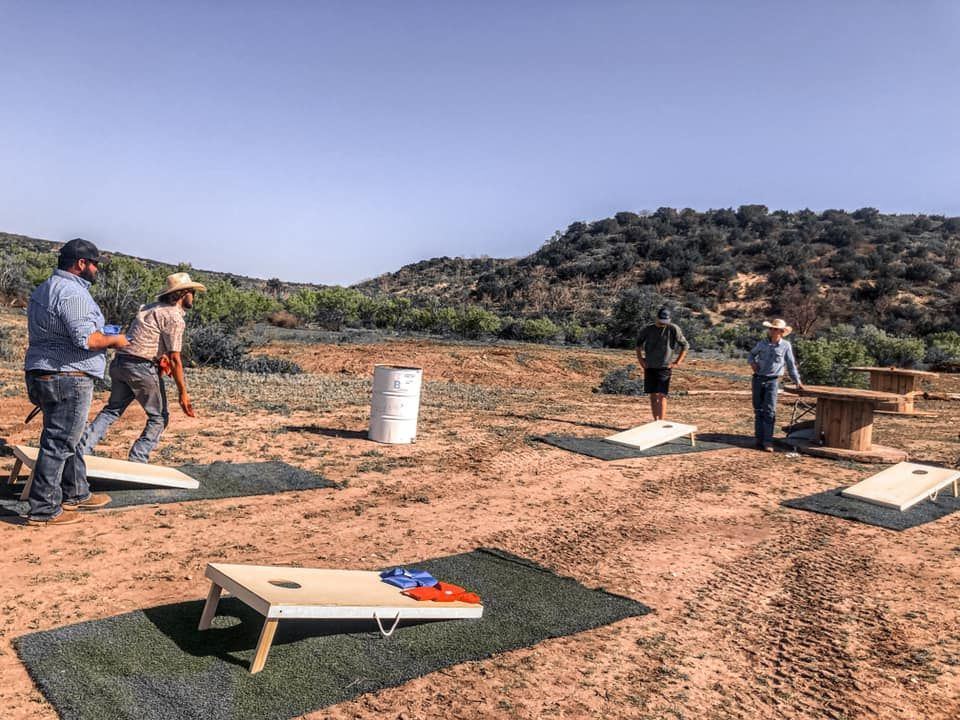 People playing outdoor cornhole on a dirt field with hills in the background
