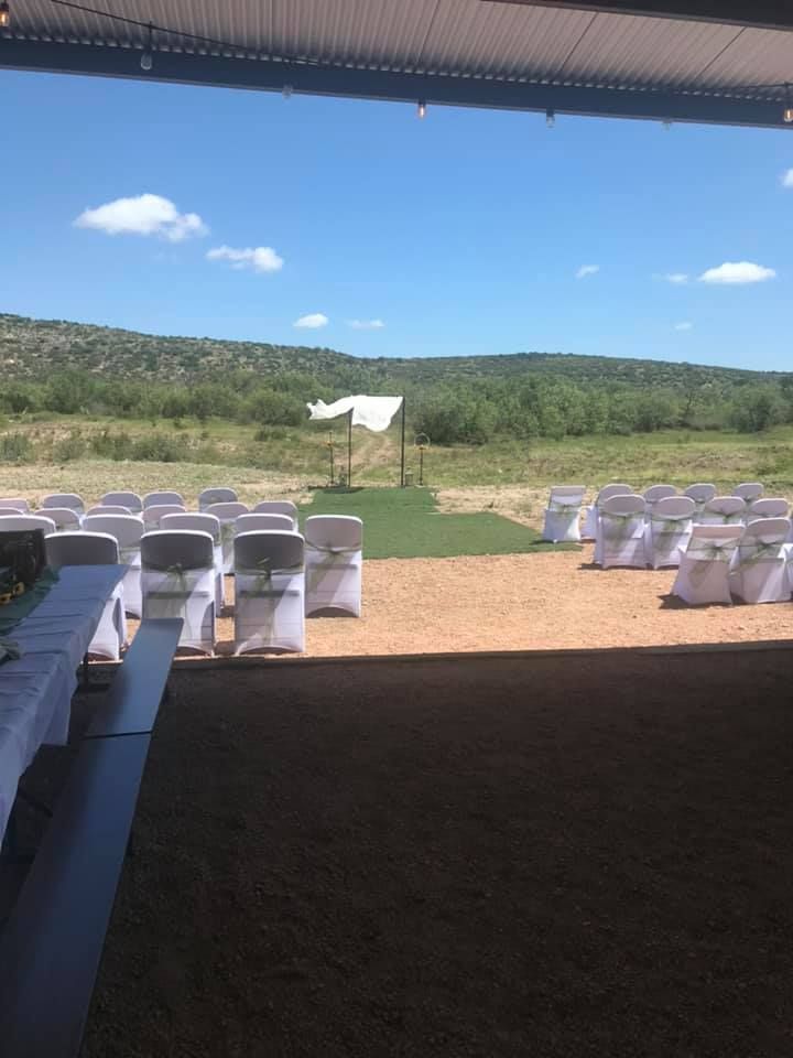 Outdoor ceremony setup with white chairs under a canopy, overlooking green hills and a blue sky
