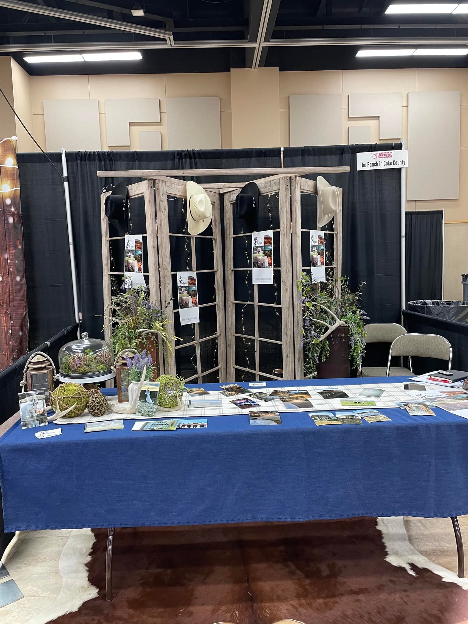 Booth display with blue tablecloth, clear acrylic panels, plants, and papers in an indoor exhibition hall